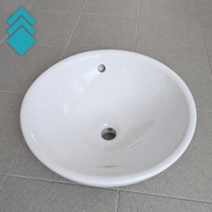 White oval ceramic sink basin on gray tiled floor, with drain hole and small overflow hole near the rim; blue stacked-diamond logo in the top-left corner.